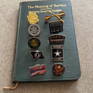 May include: A green book titled "The Meaning of Service" by Harry Emerson Fosdick with a collection of military pins and badges attached to the cover. The pins include the National Museum of the United States Army, a Korean War Veteran pin, a U.S. Army Veteran pin, and a pin with the text "CT Veteran 2009-2010".