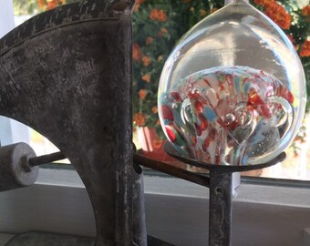 May include: A vintage metal scale with a glass orb filled with colorful glass swirls. The orb is suspended from the scale's arm and sits on a metal platform. The scale is resting on a wooden surface.