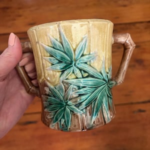 May include: A ceramic mug with a bamboo-style handle and body. The mug is tan with vertical lines and features green and blue-green leaf designs. The handles are brown, and the mug is held by a hand.