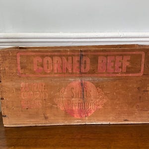 May include: A vintage wooden crate for corned beef, with red lettering that reads "CORNED BEEF" and "Swift's Premium". The crate is brown and has a worn, rustic appearance.
