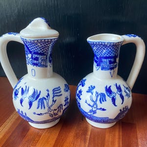 May include: Two white ceramic pitchers with blue willow pattern designs. One pitcher has a lid. Both have curved handles and are decorated with birds, trees, and architectural elements. The pitchers are on a wooden surface.