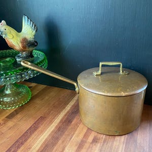 May include: A vintage copper saucepan with a lid and a long handle. The pot has a brass handle on the lid. A decorative ceramic rooster and a green glass cake stand are in the background.