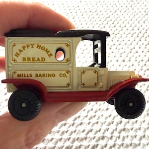 May include: A vintage toy delivery truck with the words "Happy Home Bread" and "Mills Baking Co." printed on the side. The truck is cream-colored with a black roof and red accents. The toy is held in a hand.