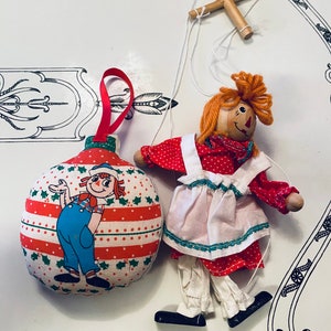 May include: A red and green Christmas ornament shaped like a ball with a cartoon image of a girl in a red dress and blue overalls. The ornament is attached to a string and a wooden puppet of a girl in a red dress and white apron.