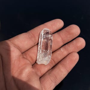 Puede incluir: Una punta de cristal de cuarzo transparente y alargada con estructura hexagonal y facetas naturales. El espécimen mineral es translúcido con inclusiones internas visibles y una terminación puntiaguda, mostrando una textura superficial rugosa.