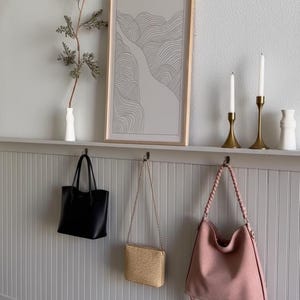 Pu&ograve; includere: Una scena d'interni minimalista presenta una borsa tote nera, una borsa di paglia intrecciata e una borsa a tracolla rosa appese a dei ganci. Una stampa artistica astratta incorniciata, candele e un vaso con vegetazione sono esposti su una mensola.