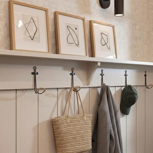 Pu&ograve; includere: Un ingresso dai toni neutri con arte astratta incorniciata, una lampada da parete e una fila di ganci. Una borsa tote intrecciata, una giacca grigia e un berretto verde sono appesi ai ganci. Il muro &egrave; dipinto di un colore chiaro.
