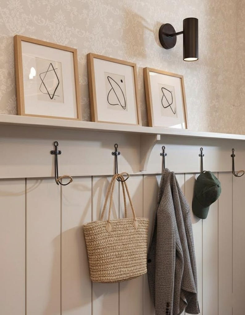 Pu&ograve; includere: Un ingresso dai toni neutri con arte astratta incorniciata, una lampada da parete e una fila di ganci. Una borsa tote intrecciata, una giacca grigia e un berretto verde sono appesi ai ganci. Il muro &egrave; dipinto di un colore chiaro.