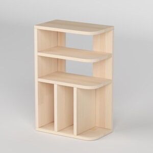 May include: A light-colored wooden bookshelf with three shelves and a vertical design. The shelves have rounded corners, and the unit has a natural wood grain finish. The bookshelf is designed for storage and display.