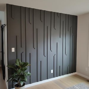 以下が含まれることがあります： 垂直の線形デザイン要素を備えた、濃い灰色のアクセントウォール。壁には、モダンでテクスチャード加工された外観を作り出す、一連の隆起した角度の付いた線があります。大きな緑の葉を持つ鉢植えの植物が壁の前にあります。