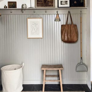 Pu&ograve; includere: Una scena d'interni accogliente con una parete grigio chiaro a pannelli adornata da opere d'arte e una mensola. Un disegno a tratto incorniciato &egrave; appeso sotto, insieme a una borsa tote in pelle, un piccolo sgabello in legno e un cesto portabiancheria in tela bianca. Un tappeto con un motivo blu e beige &egrave; sul pavimento.