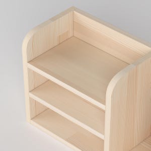 May include: A light-colored wooden shelf unit with three shelves. The unit has rounded corners and a simple, modern design. The wood grain is visible, giving it a natural look. The shelves are evenly spaced, providing storage space.