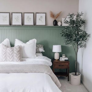 Pu&ograve; includere: Interno di una camera da letto con un letto con lenzuola bianche, cuscini decorativi e una coperta lavorata a maglia. Sopra il letto, una parete verde espone stampe botaniche incorniciate. Un comodino in legno con una lampada e un ulivo completano l'arredamento.