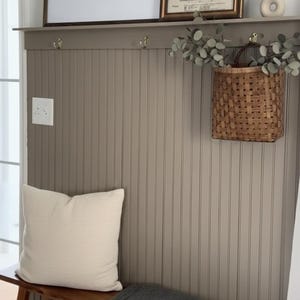Pu&ograve; includere: Scena d'interni accogliente con una panca in legno, un cuscino color crema e una coperta grigio scuro. Sopra, una foto in bianco e nero incorniciata e una stampa vintage della Torre Eiffel sono appese a una parete grigia a pannelli. Un cesto intrecciato con vegetazione aggiunge un tocco naturale.