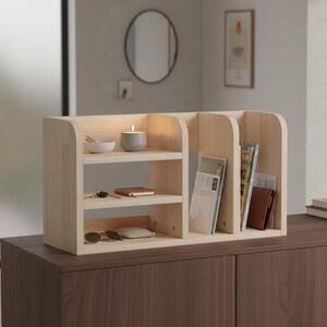 May include: A light-colored wooden desk organizer with multiple shelves and compartments. The organizer holds a small candle, a bowl, keys, a notebook, sunglasses, and books. The unit sits on a dark brown cabinet.