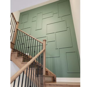 May include: A wooden staircase with black metal railings ascends towards a sage green wall. The wall features a series of rectangular and square decorative panels. The stairs have light-coloured wooden steps and a matching handrail.
