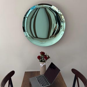 Könnte beinhalten: Ein runder, türkis gefärbter Spiegel hängt über einem Holztisch. Auf dem Tisch befinden sich ein Laptop, eine Vase mit roten Rosen und eine Computermaus. Zwei dunkle Holzstühle flankieren den Tisch und bilden einen Arbeitsbereich.