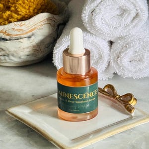 K&ouml;nnte beinhalten: Eine Glasflasche Minescence Rose Squalane Serum mit einer Pipette, die auf einer wei&szlig;en quadratischen Schale mit goldenem Rand ruht. Ein goldfarbenes schleifenf&ouml;rmiges Accessoire befindet sich neben der Flasche. Gerollte wei&szlig;e Handt&uuml;cher und ein gelber Schwamm in einer dekorativen Schale sind im Hintergrund.