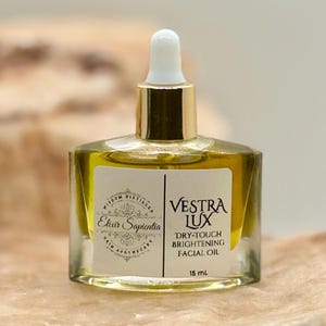 May include: A clear glass bottle of facial oil with a gold cap and white dropper. The bottle contains a yellow liquid. The label reads 'Vestra Lux Dry-Touch Brightening Facial Oil' and 'Elixir Sapientia Skin Apothecary'. The bottle sits on a light brown, textured surface.