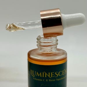K&ouml;nnte beinhalten: Eine klare Glasflasche mit einer Pipette, gef&uuml;llt mit einer goldenen Fl&uuml;ssigkeit. Die Flasche hat ein dunkelgr&uuml;nes Etikett mit dem Wort "LUMINESCEN" und "Vitamin C & Rose Squalane". Die Pipette hat eine ros&eacute;goldene Kappe und einen wei&szlig;en Aufsatz.