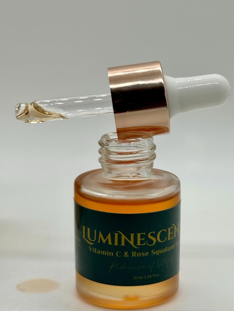 K&ouml;nnte beinhalten: Eine klare Glasflasche mit einer Pipette, gef&uuml;llt mit einer goldenen Fl&uuml;ssigkeit. Die Flasche hat ein dunkelgr&uuml;nes Etikett mit dem Wort "LUMINESCEN" und "Vitamin C & Rose Squalane". Die Pipette hat eine ros&eacute;goldene Kappe und einen wei&szlig;en Aufsatz.