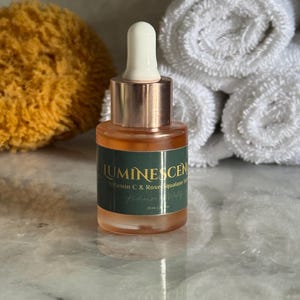 K&ouml;nnte beinhalten: Kleine Glasflasche mit Serum, mit wei&szlig;er Tropferspitze und kupferfarbenem Hals. Die Flasche enth&auml;lt eine klare, orangefarbene Fl&uuml;ssigkeit und hat ein gr&uuml;nes Etikett mit dem Text "LUMINESCEN Vitamin C & Rose Squalane Serum". Die Flasche ist 20 ml.