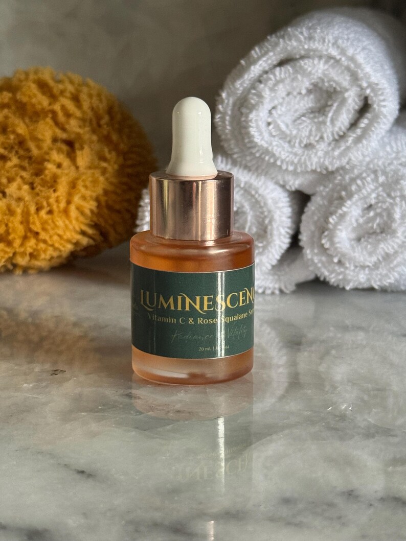 K&ouml;nnte beinhalten: Kleine Glasflasche mit Serum, mit wei&szlig;er Tropferspitze und kupferfarbenem Hals. Die Flasche enth&auml;lt eine klare, orangefarbene Fl&uuml;ssigkeit und hat ein gr&uuml;nes Etikett mit dem Text "LUMINESCEN Vitamin C & Rose Squalane Serum". Die Flasche ist 20 ml.
