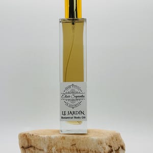 May include: A rectangular glass bottle of "LE JARDIN Botanical Body Oil" with a gold cap, resting on a light brown stone. The bottle contains a golden-colored liquid and has a white label with black text.