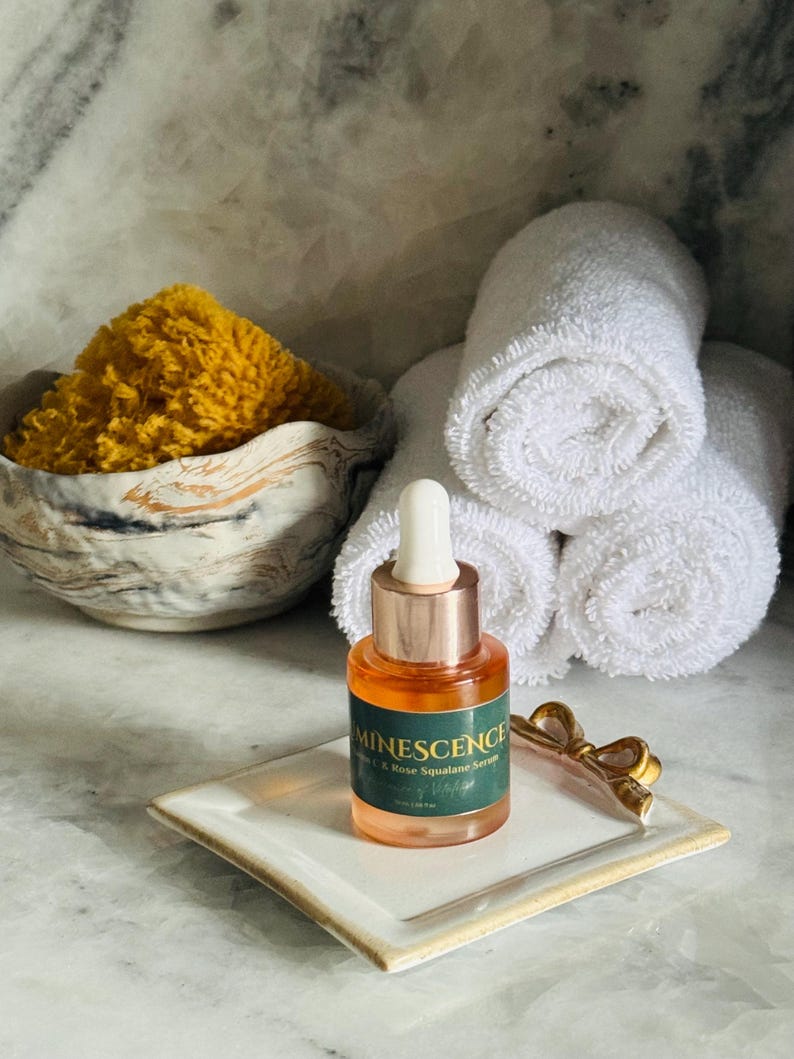 K&ouml;nnte beinhalten: Eine Glasflasche Minescence Rose Squalane Serum mit einer Pipette, die auf einer wei&szlig;en quadratischen Schale mit goldenem Rand ruht. Ein goldfarbenes schleifenf&ouml;rmiges Accessoire befindet sich neben der Flasche. Gerollte wei&szlig;e Handt&uuml;cher und ein gelber Schwamm in einer dekorativen Schale sind im Hintergrund.
