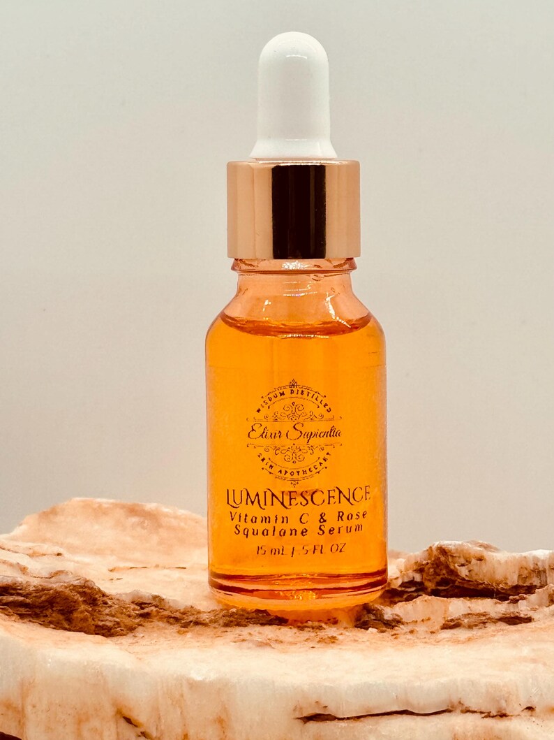 K&ouml;nnte beinhalten: Eine Glasflasche mit Luminescence Vitamin C & Rose Squalane Serum. Die Flasche hat eine Tropferspitze mit einem wei&szlig;en Kolben und einem ros&eacute;goldenen Kragen. Das Serum ist orange und das Etikett lautet "Elixir Sapientia". Die Flasche ist 15 ml.
