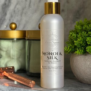 May include: A pearlescent bottle of "MONOI & SILK" apothecary shaving oil for women, with a gold pump and cap. The bottle is 5 fl oz. A rose gold razor and a small green plant are also visible.