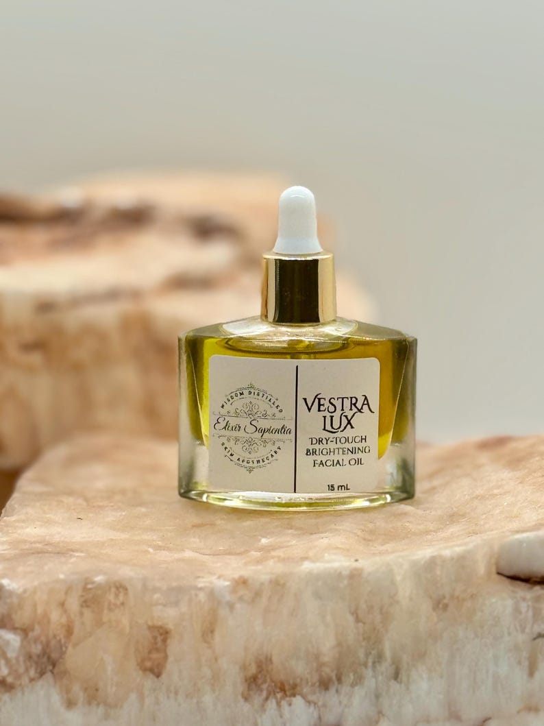 May include: A clear glass bottle of facial oil with a gold cap and white dropper. The bottle contains a yellow liquid. The label reads 'Vestra Lux Dry-Touch Brightening Facial Oil' and 'Elixir Sapientia Skin Apothecary'. The bottle sits on a light brown, textured surface.