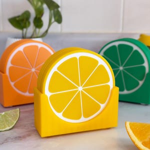 Könnte beinhalten: Ein Set von Untersetzern im Zitrus-Design in Orange, Gelb und Grün, jeweils mit einem weißen Segmentmuster. Die Untersetzer werden in passenden Haltern präsentiert. Eine Orangenscheibe und eine Limette befinden sich im Vordergrund.