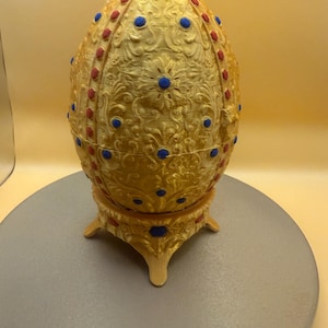 May include: A golden egg-shaped decorative object with ornate floral patterns and red and blue accents. The egg sits on a matching stand with three legs, creating a regal and elegant appearance. This decorative piece is ideal for display.