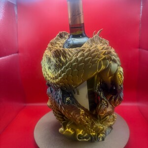 Peut inclure: Une bouteille de vin dans un support décoratif texturé de couleur or. Le support présente un motif en forme de dragon, avec des écailles et des détails complexes. La bouteille a un col doré et une étiquette avec du texte.