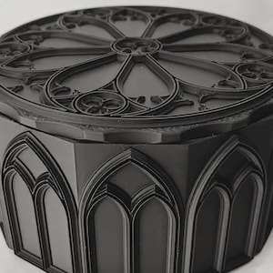 May include: A matte black circular storage box featuring a lid with a raised rose window pattern. The sides of the container are decorated with a series of repeating arched window motifs, creating a structured, multi-faceted exterior design.