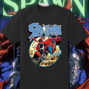 May include: Black t-shirt featuring a graphic of the comic book character Spawn. The design includes the word "SPAWN" in blue letters with a skull and chain detail. The character is depicted in a dynamic pose with a red cape.