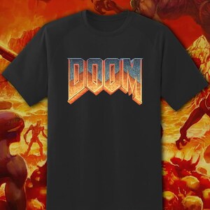 May include: Black t-shirt featuring the word "DOOM" in a 3D, textured design with orange and blue hues. The shirt is set against a fiery, hellish background with demonic figures, evoking a video game theme.