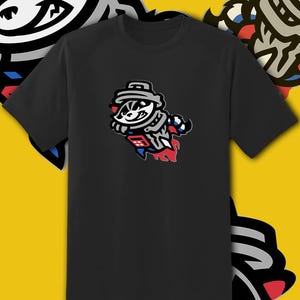 May include: Black t-shirt featuring a cartoon badger mascot in a rocket ship design. The badger is wearing a hat and a jersey with red, white, and blue accents. The design is a stylized illustration.