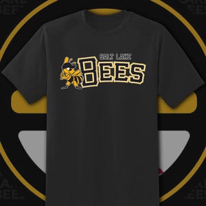 May include: Black t-shirt featuring the words "SALT LAKE BEES" in gold and white lettering. A cartoon bee holding a baseball bat is on the left. The shirt has a crew neck and short sleeves.