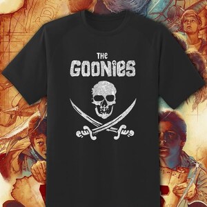 May include: Black t-shirt featuring a white graphic of a skull and crossed swords, with the text "THE GOONIES" above. The design has a distressed, vintage look. The shirt is a classic crew neck.