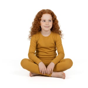 May include: A young person with long, curly red hair sits cross-legged, wearing a mustard-yellow long-sleeved shirt and matching pants. The outfit is a solid color, and the person is looking slightly to the side.