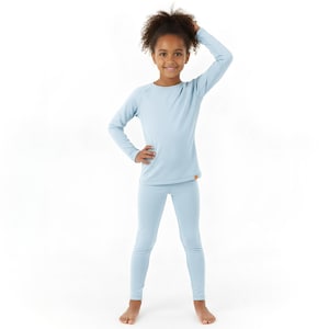 May include: A young person wearing a light blue long-sleeved shirt and matching leggings. The outfit is a solid color and appears to be made of a soft, comfortable material. The person is smiling and posing for the camera.