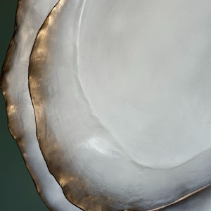 May include: Close-up of two white, organically shaped plates with a metallic gold rim. The plates have a textured surface and are stacked, creating a layered effect. The background is a muted green.