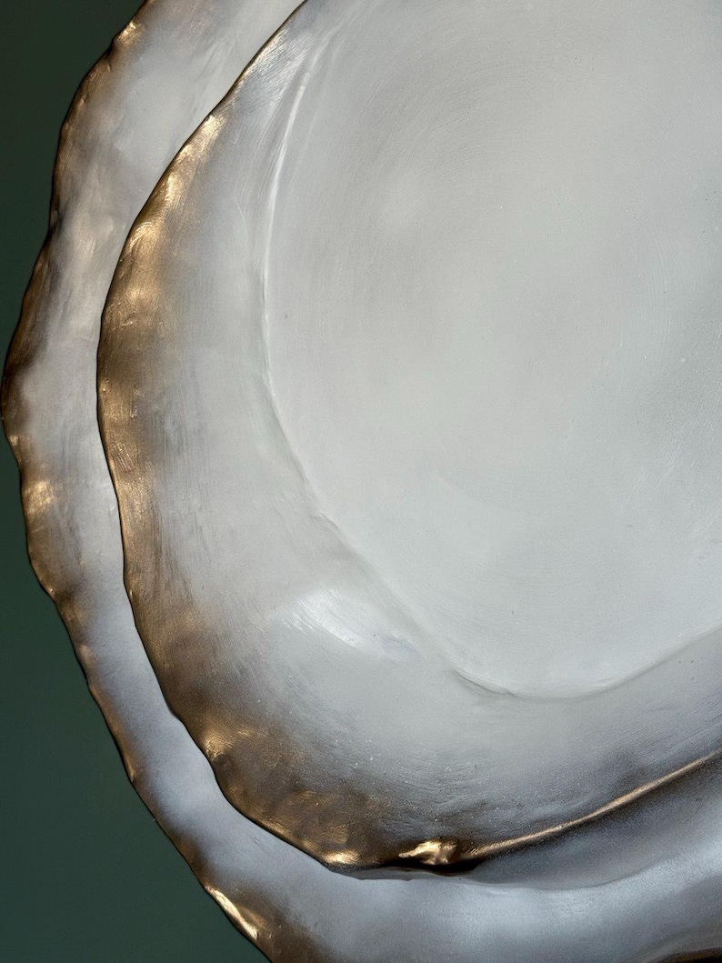 May include: Close-up of two white, organically shaped plates with a metallic gold rim. The plates have a textured surface and are stacked, creating a layered effect. The background is a muted green.
