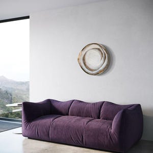 May include: A modern living room featuring a plush, deep purple sofa with a low profile. A decorative, circular wall art piece with a swirling design in neutral tones hangs above the sofa. The room has a minimalist design with a large window.