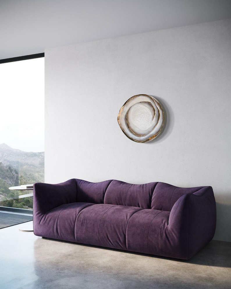 May include: A modern living room featuring a plush, deep purple sofa with a low profile. A decorative, circular wall art piece with a swirling design in neutral tones hangs above the sofa. The room has a minimalist design with a large window.