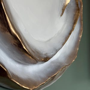 May include: A decorative, abstract ceramic bowl with a white interior and gold-colored edges. The bowl has a textured surface and a unique, organic shape. The gold trim adds a touch of elegance to the piece. The bowl is likely used for decorative purposes.