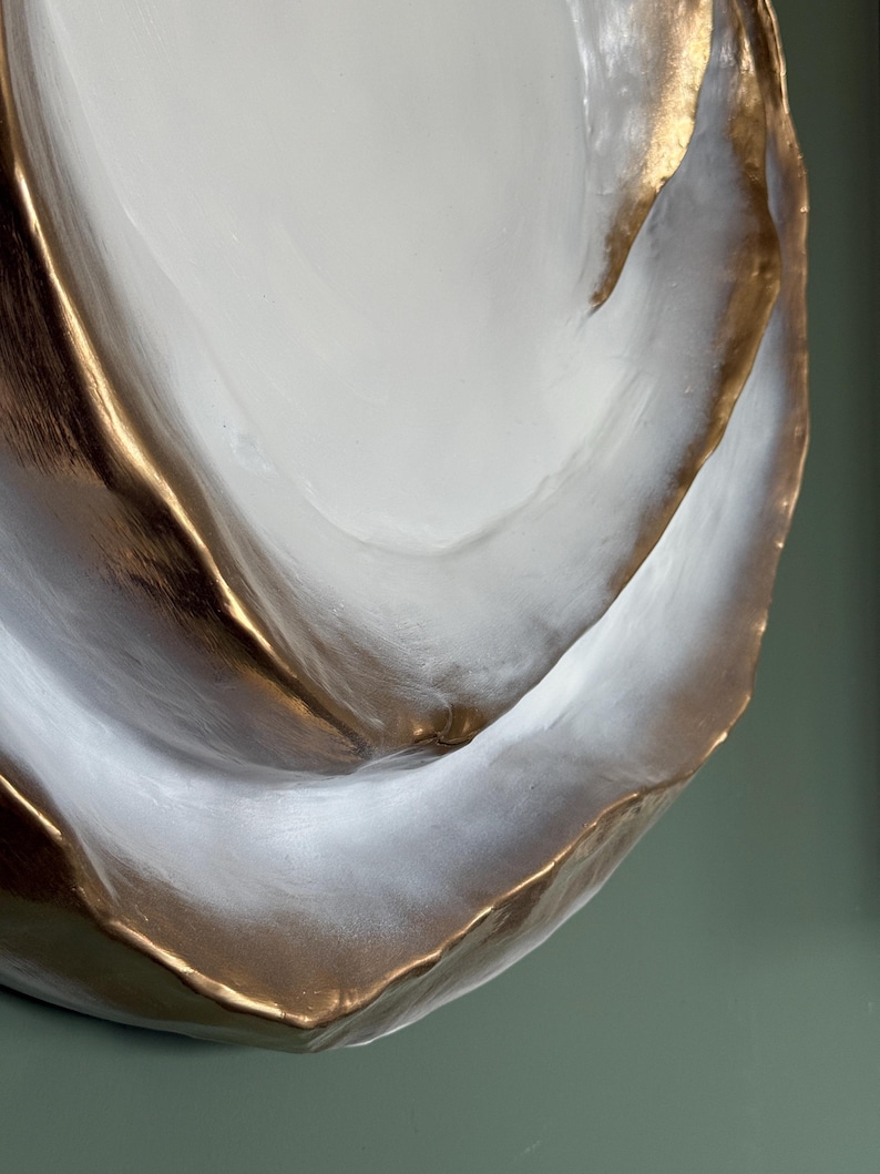May include: A decorative, abstract ceramic bowl with a white interior and gold-colored edges. The bowl has a textured surface and a unique, organic shape. The gold trim adds a touch of elegance to the piece. The bowl is likely used for decorative purposes.