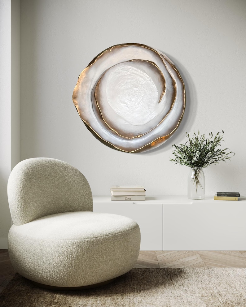 May include: A large, circular wall art piece with a swirling design. The artwork features a white center, surrounded by layers of white and gold, creating a textured, abstract appearance. The piece is mounted on a white wall, above a white cabinet.
